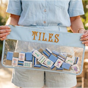 May include: A clear pouch with a light blue top and gold zip, embroidered with the word "TILES". The pouch contains a collection of blue, white, and tan mahjong tiles. The tiles feature various symbols and numbers.