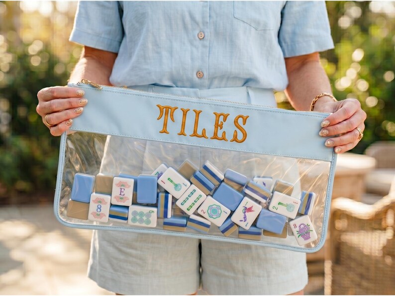 May include: A clear pouch with a light blue top and gold zip, embroidered with the word "TILES". The pouch contains a collection of blue, white, and tan mahjong tiles. The tiles feature various symbols and numbers.