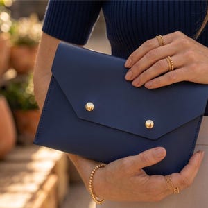 May include: A navy blue envelope clutch with two gold button fastenings. The clutch is held by a person wearing gold jewellery, including rings and a bracelet. The background features terracotta plant pots and greenery.