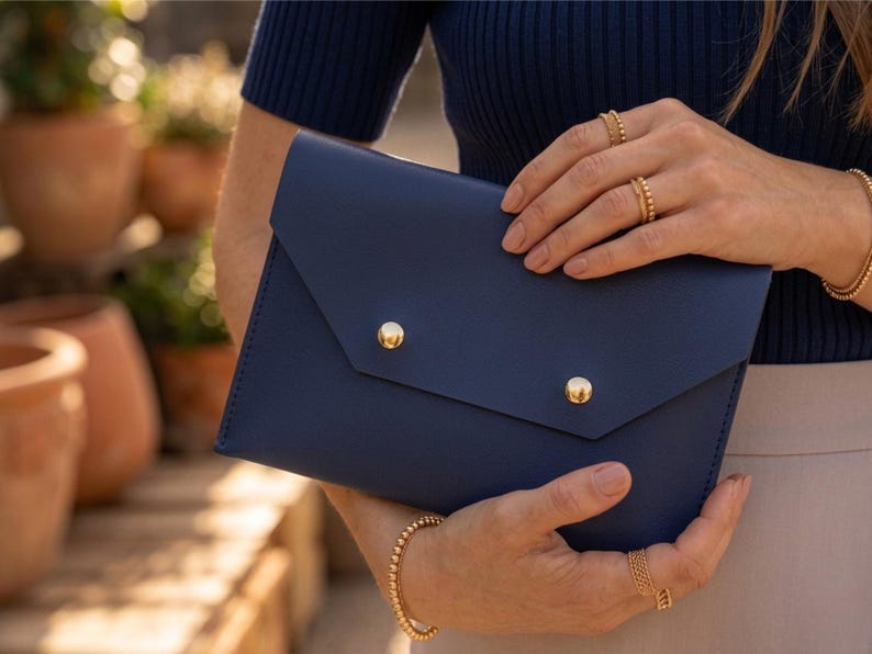 May include: A navy blue envelope clutch with two gold button fastenings. The clutch is held by a person wearing gold jewellery, including rings and a bracelet. The background features terracotta plant pots and greenery.
