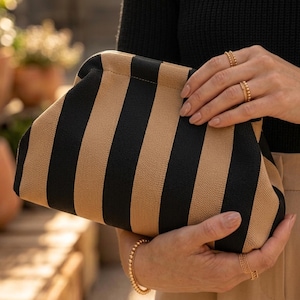 Pode incluir: Uma bolsa clutch estruturada com um padr&atilde;o de riscas verticais arrojado em cores alternadas preto e bege. A bolsa tem um exterior de tecido texturizado macio com um fecho de moldura curva, segurada por duas m&atilde;os com an&eacute;is dourados.