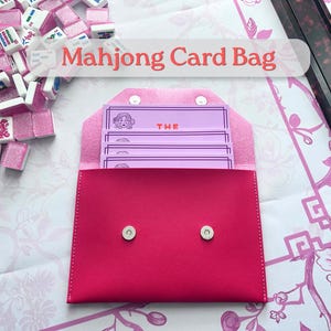 May include: A vibrant pink Mahjong card bag with a snap closure, containing several light purple cards. The cards feature a woman's face and the word "THE". Mahjong tiles are scattered around the bag.