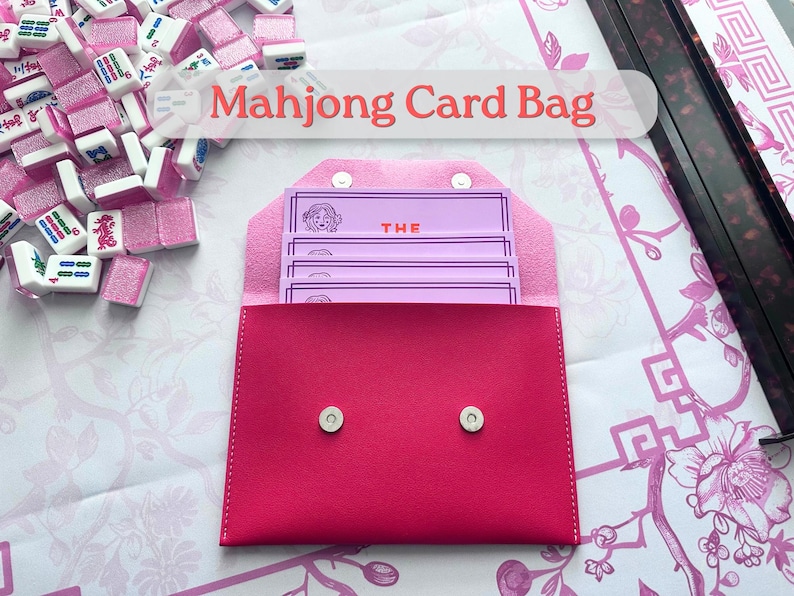 May include: A vibrant pink Mahjong card bag with a snap closure, containing several light purple cards. The cards feature a woman's face and the word "THE". Mahjong tiles are scattered around the bag.