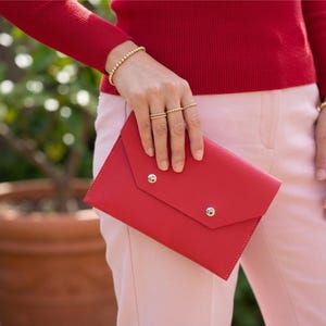 May include: A red leather clutch with two silver press studs. The clutch is held by a person wearing a red jumper, gold beaded bracelets, and gold rings. The person is also wearing pink trousers.