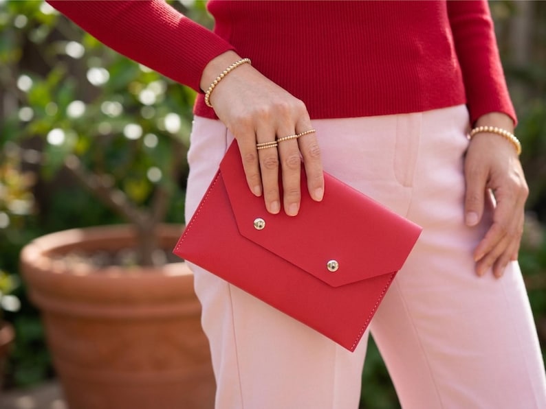 May include: A red leather clutch with two silver press studs. The clutch is held by a person wearing a red jumper, gold beaded bracelets, and gold rings. The person is also wearing pink trousers.