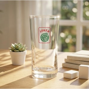 May include: A clear glass tumbler with a red and green "JOKER" logo. The glass is on a wooden table next to a small potted succulent and two white rectangular objects. A stack of books is in the background.