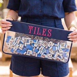 May include: A navy blue zipped pouch with the word "TILES" embroidered in pink. The clear front reveals a collection of white and blue Mahjong tiles. The pouch is held by a person wearing a navy blue outfit.