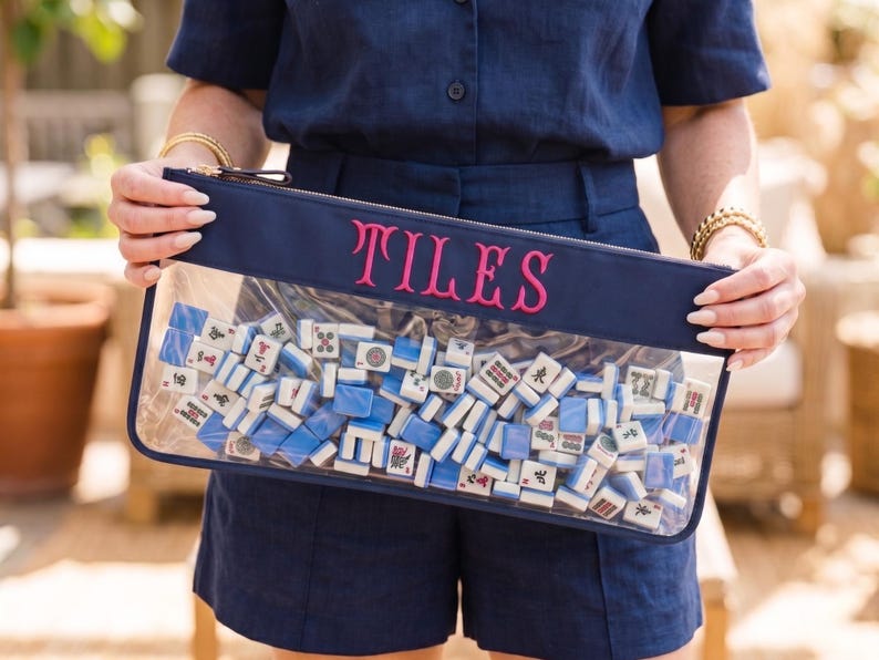 May include: A navy blue zipped pouch with the word "TILES" embroidered in pink. The clear front reveals a collection of white and blue Mahjong tiles. The pouch is held by a person wearing a navy blue outfit.