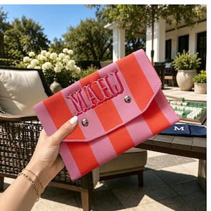 May include: A rectangular fabric clutch featuring vertical stripes in bright orange and light pink. The front flap is secured with two silver-toned metal snaps and displays the embroidered monogram letters MAHJ in a dark pink thread.