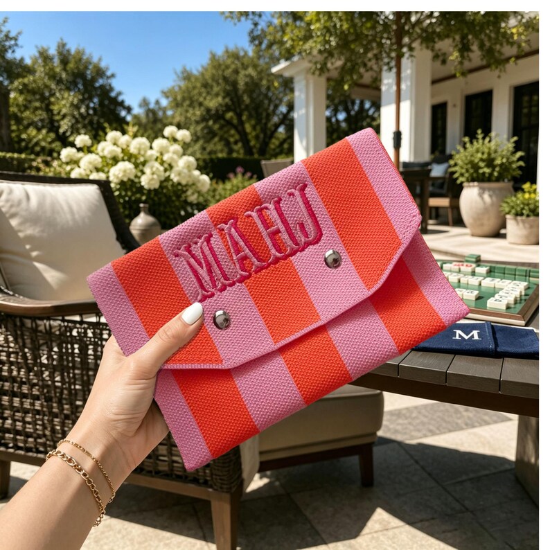 May include: A rectangular fabric clutch featuring vertical stripes in bright orange and light pink. The front flap is secured with two silver-toned metal snaps and displays the embroidered monogram letters MAHJ in a dark pink thread.