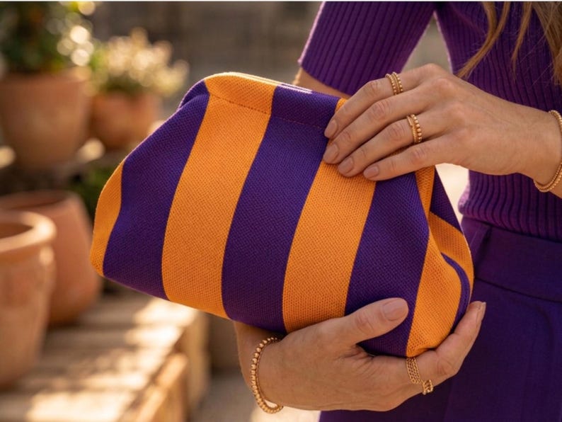 Pode incluir: Uma bolsa clutch com um padr&atilde;o ousado de listras verticais em tons alternados de roxo profundo e laranja vibrante. A bolsa tem uma textura semelhante a tecido e uma estrutura arredondada que &eacute; segurada firmemente por duas m&atilde;os.