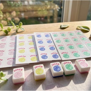 May include: A collection of Mahjong Joker tiles and sticker sheets. The tiles are white with colored designs and the word "JOKER". Sticker sheets feature the word "JOKER" and matching colored designs. The color palette includes pink, yellow, blue, and green.