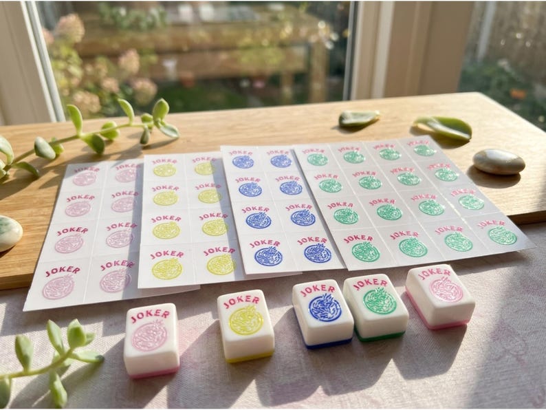 May include: A collection of Mahjong Joker tiles and sticker sheets. The tiles are white with colored designs and the word "JOKER". Sticker sheets feature the word "JOKER" and matching colored designs. The color palette includes pink, yellow, blue, and green.