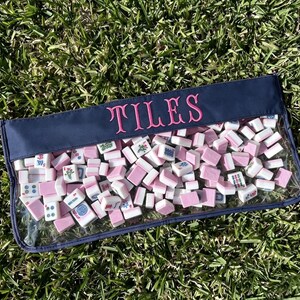 May include: A clear pouch filled with pink and white mahjong tiles, resting on green grass. The pouch has a navy blue top with the word "TILES" embroidered in pink. The tiles are a mix of shapes and designs.