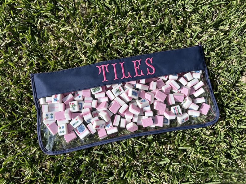 May include: A clear pouch filled with pink and white mahjong tiles, resting on green grass. The pouch has a navy blue top with the word "TILES" embroidered in pink. The tiles are a mix of shapes and designs.