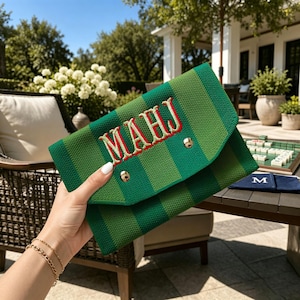 May include: A green and dark green striped fabric clutch bag featuring the embroidered letters MAHJ in red with a white outline. The envelope-style bag has a pointed flap secured by two gold-toned metal snap buttons on the front exterior.