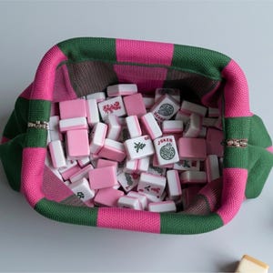 May include: A knitted pouch with a checkered pattern in dark green and bright pink, filled with a collection of rectangular mahjong tiles. The tiles are white with pink backs and feature various black and red symbols, including one marked Joker.