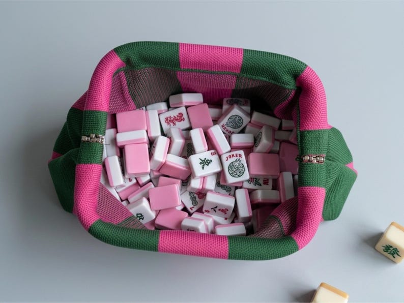 May include: A knitted pouch with a checkered pattern in dark green and bright pink, filled with a collection of rectangular mahjong tiles. The tiles are white with pink backs and feature various black and red symbols, including one marked Joker.