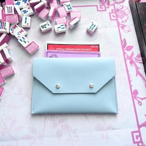 May include: A pale blue Mahjong tile case with silver press studs, containing rule cards, surrounded by pink and white Mahjong tiles. The tiles are scattered on a pink and white patterned surface.