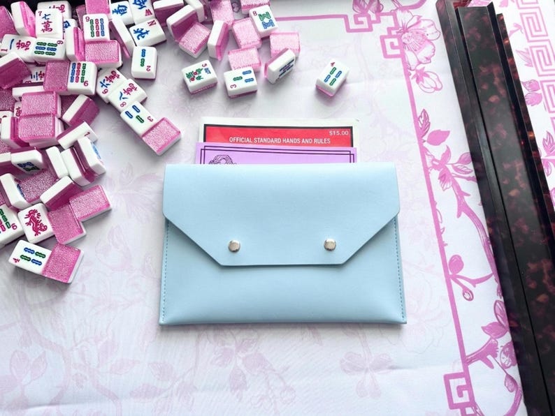 May include: A pale blue Mahjong tile case with silver press studs, containing rule cards, surrounded by pink and white Mahjong tiles. The tiles are scattered on a pink and white patterned surface.