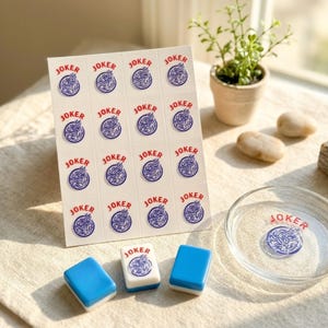 May include: A collection of game pieces and accessories featuring a "JOKER" theme. Includes a sheet of stickers, game tiles, and a small glass dish, all with a matching design in blue and red. A small potted plant and decorative stones are also present.