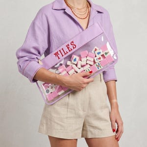 May include: A clear rectangular pouch with the word "TILES" in red embroidery. The pouch is filled with pink and white Mahjong tiles. The pouch has a lavender border.