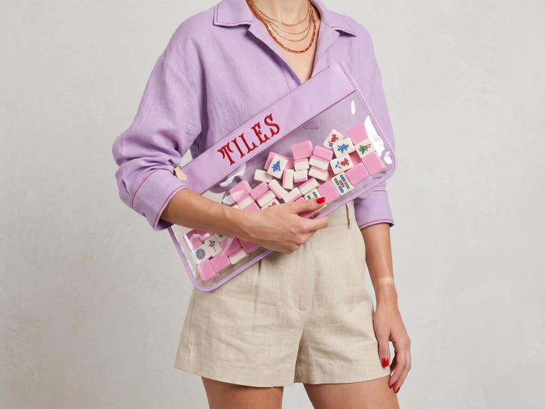 May include: A clear rectangular pouch with the word "TILES" in red embroidery. The pouch is filled with pink and white Mahjong tiles. The pouch has a lavender border.