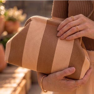 Pode incluir: Uma bolsa clutch estruturada com um padr&atilde;o de riscas verticais em tons alternados de bege e castanho. A bolsa tem um fecho de moldura superior curvo e um exterior de tecido texturado, segurada por duas m&atilde;os adornadas com an&eacute;is e pulseiras de contas douradas.