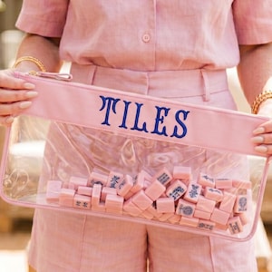 May include: A clear rectangular pouch with a pink top and the word "TILES" in navy blue lettering. The pouch is filled with pink mahjong tiles. The person in the image is wearing a pink shirt and shorts.