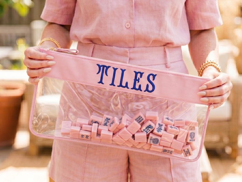May include: A clear rectangular pouch with a pink top and the word "TILES" in navy blue lettering. The pouch is filled with pink mahjong tiles. The person in the image is wearing a pink shirt and shorts.
