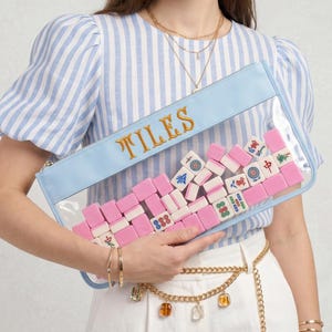 May include: A clear rectangular clutch with a pale blue border and the word "TILES" in gold lettering. The clutch is filled with pink and white Mahjong tiles. The clutch is held by a person wearing a blue and white striped top.