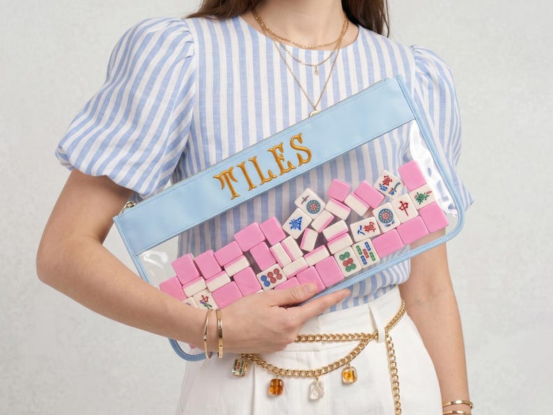 May include: A clear rectangular clutch with a pale blue border and the word "TILES" in gold lettering. The clutch is filled with pink and white Mahjong tiles. The clutch is held by a person wearing a blue and white striped top.