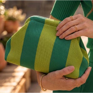 Pode incluir: Uma bolsa clutch estruturada com um padr&atilde;o de riscas verticais arrojado em tons alternados de verde floresta e verde lima. A bolsa tem uma textura macia semelhante a malha e um fecho superior curvo em estilo de moldura.