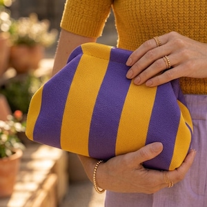 May include: A knit clutch bag featuring vertical stripes in vibrant purple and mustard yellow. The bag has a soft, rounded frame construction with a hidden closure mechanism, showcasing a textured fabric surface and a structured, pouch-like shape.