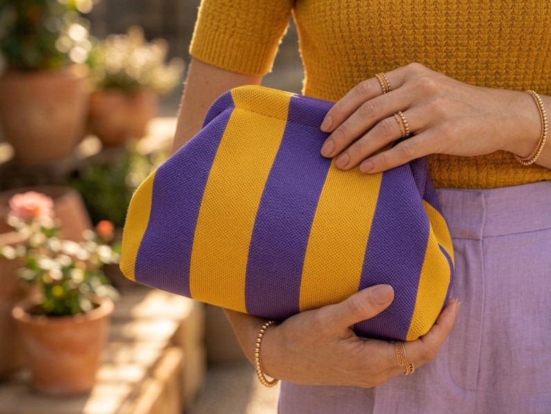 May include: A knit clutch bag featuring vertical stripes in vibrant purple and mustard yellow. The bag has a soft, rounded frame construction with a hidden closure mechanism, showcasing a textured fabric surface and a structured, pouch-like shape.
