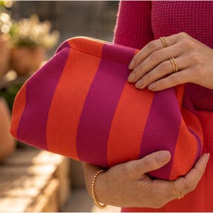 May include: A clutch bag featuring a vibrant pattern of alternating vertical stripes in bright orange and deep magenta. The bag has a soft, textured knit construction with a rounded, frame-style opening that closes securely at the top.
