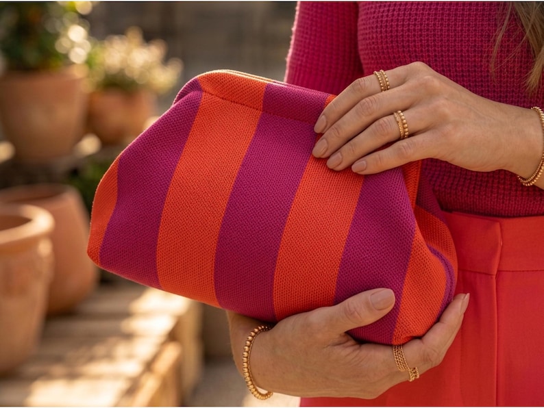 May include: A clutch bag featuring a vibrant pattern of alternating vertical stripes in bright orange and deep magenta. The bag has a soft, textured knit construction with a rounded, frame-style opening that closes securely at the top.
