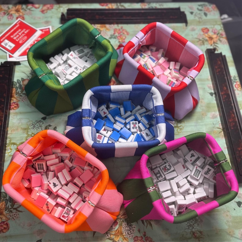 May include: Five fabric pouches with striped patterns in green, red, blue, orange, and pink, each filled with small white, blue, or pink mahjong tiles. The pouches feature a structured, open-top design with a metal frame closure at the top edge.