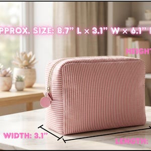 May include: Pink and white striped cosmetic bag, approximately 8.7" long, 3.1" wide, and 6.1" high. The bag has a zipper closure and a small pink charm. The bag is sitting on a white surface.