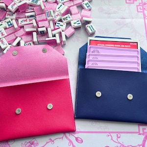 May include: A close-up of two leather pouches, one bright pink and one navy blue, each with silver snap closures. The pink pouch is open, revealing a lighter pink interior. The navy pouch contains pink cards. Mahjong tiles are scattered around.