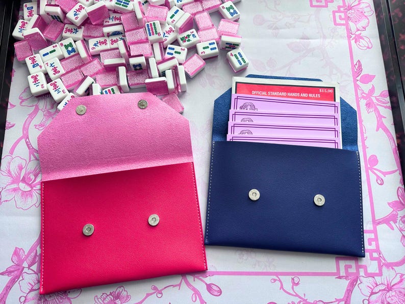May include: A close-up of two leather pouches, one bright pink and one navy blue, each with silver snap closures. The pink pouch is open, revealing a lighter pink interior. The navy pouch contains pink cards. Mahjong tiles are scattered around.