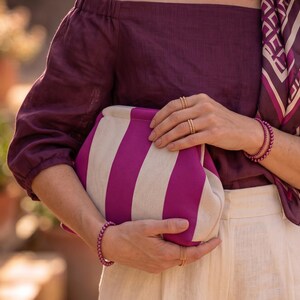 Pode incluir: Uma bolsa clutch com largas riscas verticais em magenta e branco sujo. A bolsa &eacute; segurada contra um top roxo escuro de ombros desca&iacute;dos e cal&ccedil;as claras. O acess&oacute;rio &eacute; combinado com an&eacute;is e pulseiras de contas em tom dourado.