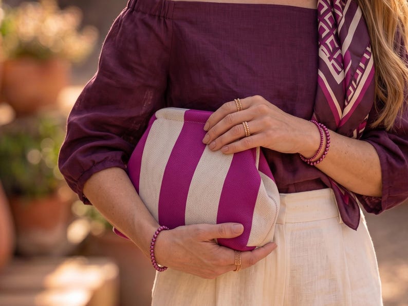 Pode incluir: Uma bolsa clutch com largas riscas verticais em magenta e branco sujo. A bolsa &eacute; segurada contra um top roxo escuro de ombros desca&iacute;dos e cal&ccedil;as claras. O acess&oacute;rio &eacute; combinado com an&eacute;is e pulseiras de contas em tom dourado.