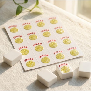 May include: A close-up of a set of Mahjong tiles and Joker stickers. The white tiles are rectangular, and the stickers are white with a yellow design and the word "JOKER" in red. The items are on a white surface.