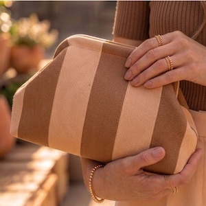 May include: A structured clutch bag featuring a vertical striped pattern in alternating shades of beige and brown. The bag has a curved top frame closure and a textured fabric exterior, held in place by two hands adorned with gold beaded rings and bracelets.