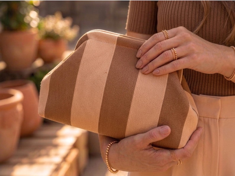 May include: A structured clutch bag featuring a vertical striped pattern in alternating shades of beige and brown. The bag has a curved top frame closure and a textured fabric exterior, held in place by two hands adorned with gold beaded rings and bracelets.