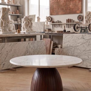 Puede incluir: Una mesa redonda con tapa de mármol y base de madera marrón oscuro con estrías. La mesa está en un taller con losas de mármol y esculturas al fondo. Un letrero de cobre en la pared dice "Chesta Crafts - Agra - Est. 1978."