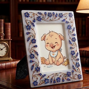Puede incluir: Un marco de fotos blanco con un diseño floral azul y dorado. El marco contiene una ilustración de un cerdito sonriente con un babero. El marco está sostenido por un soporte negro.