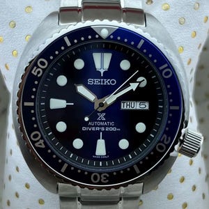 Peut inclure: Une montre de plongée automatique Seiko en acier inoxydable avec un cadran et une lunette bleus. La montre présente des marqueurs luminescents blancs, un affichage jour-date et un bracelet en acier inoxydable assorti. Le texte "AUTOMATIC DIVER'S 200m" est visible.