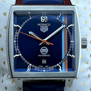 May include: A square Monaco TAG Heuer watch with a navy blue dial and a blue leather strap. The dial features white hands, orange and blue stripes, and the Gulf logo. The watch is displayed on a white dotted surface.